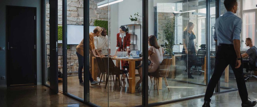 Four colleagues conducting a meeting in a glass room, as other colleagues walk past outside