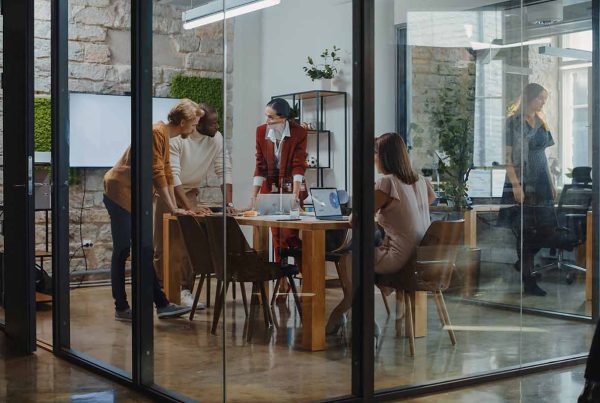 Four colleagues conducting a meeting in a glass room, as other colleagues walk past outside