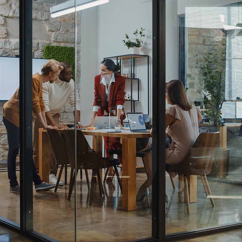 Four colleagues conducting a meeting in a glass room, as other colleagues walk past outside